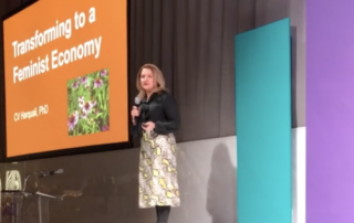 CV on stage at the SHEeo Summit, Blond lady in black top and green print skirt, holding mic