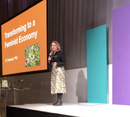 CV on stage at the SHEeo Summit, Blond lady in black top and green print skirt, holding mic